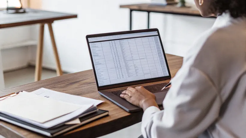 A forensic investigator seated at a desk reviewing detailed financial transaction records and account data on a laptop in a professional office setting